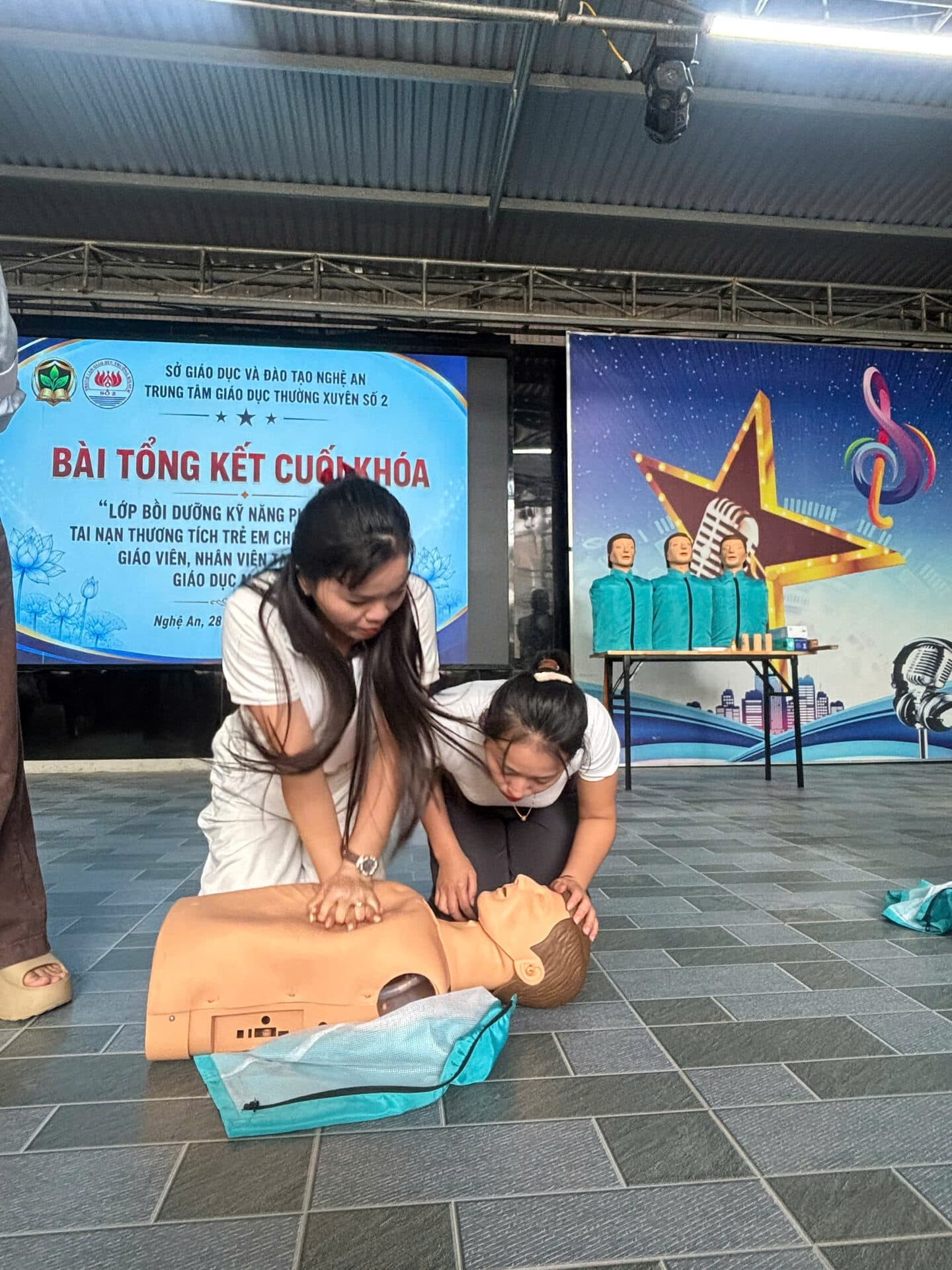 Hai giáo viên phối hợp thực hành hô hấp nhân tạo và ép tim trên mô hình trước màn hình banner buổi tổng kết cuối khóa